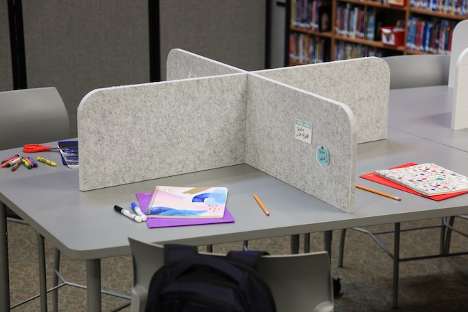 Gray felt desk privacy dividers arranged in a cross configuration on a classroom or library table, creating individual workspaces with school supplies, notebooks, and pencils visible on each side.
