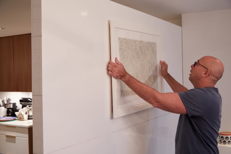 Man hanging framed artwork on a white modular partition wall in a residential interior.