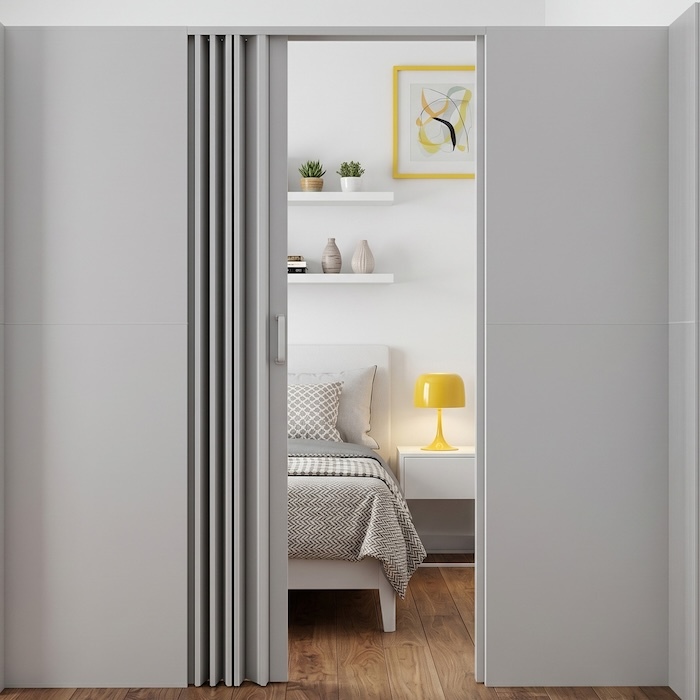 Modern minimalist bedroom partially revealed through a sliding accordion-style partition, featuring a neatly made bed, white bedside table with a yellow lamp, floating shelves with small plants and decor, and a framed abstract artwork on a light-colored wall.