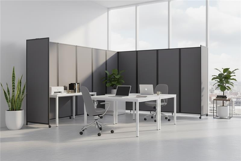 Modern office workspace with two white desks, rolling chairs, laptops, and gray privacy panels near large windows and indoor plants.