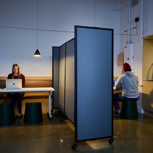 Shared indoor workspace with two individuals seated at separate desks on either side of a wheeled acoustic divider panel, featuring simple wooden benches, pendant lighting, and a clean, modern interior design.