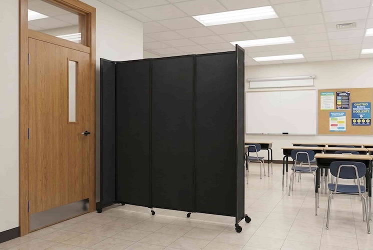 Black freestanding folding security partition on casters positioned beside a classroom doorway, with desks, chairs, a whiteboard, and bulletin board visible in the background.