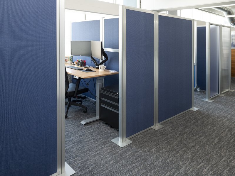 Office cubicle with a desk, monitor, chair, and blue fabric privacy panels in a rowed workspace.