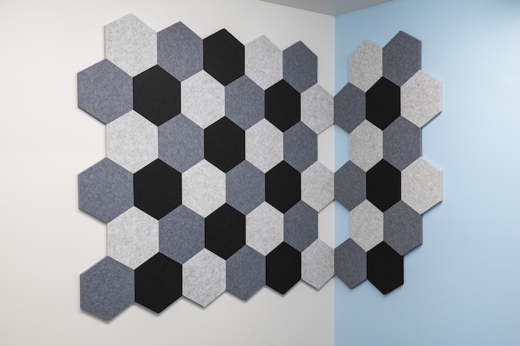 Decorative acoustic wall tiles arranged in an overlapping honeycomb pattern on a two-tone white and light blue wall. The hexagonal tiles feature a mix of light gray, dark gray, and black colors, creating a modern geometric design.