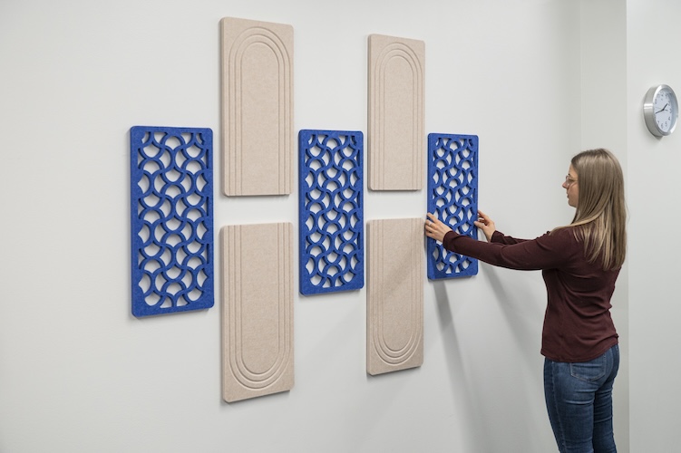 Woman arranging blue geometric SoundSorb acoustic wall tiles alongside beige arch-patterned panels on an office wall.