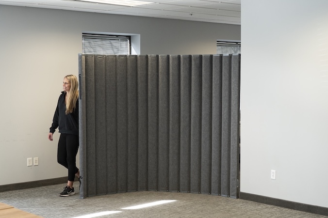 Gray freestanding acoustic room divider with a pleated design used to create a private workspace in a modern office, shown with a person walking beside the partition to demonstrate its size and mobility.