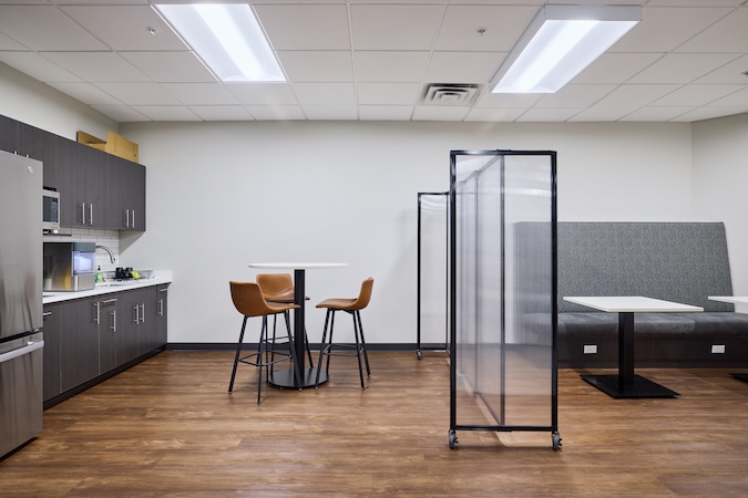 Tall transparent portable room divider separating seating areas in a modern office break room.